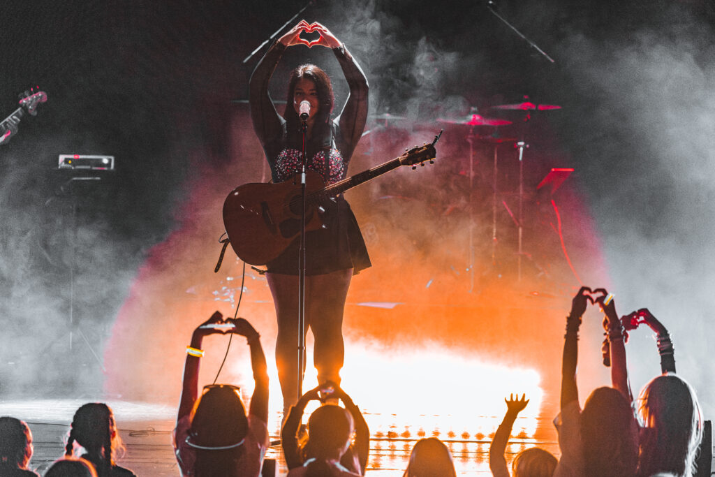 A woman standing on stage with her hands in the shape of a heart.  The audience mimicking the hearts with their hands as well.
